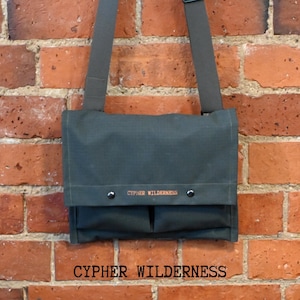 May include: A dark green messenger bag with a flap closure and two front pockets. The bag has a shoulder strap and is hanging against a red brick wall. The text "CYPHER WILDERNESS" is printed in orange on the front.
