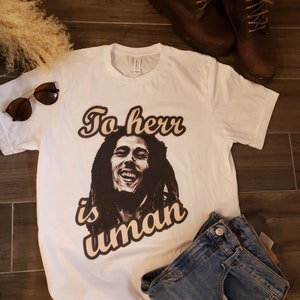 May include: White t-shirt with a black and white portrait of a smiling man with long hair, and the text "To err is human" in a brown, retro font. The shirt is styled with blue jeans, brown sunglasses, and brown leather boots.