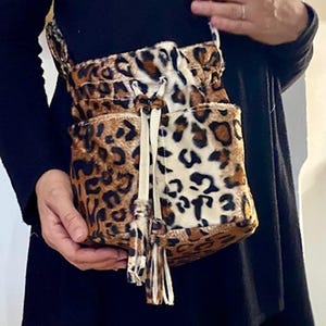 Peut inclure: Un sac à bandoulière à imprimé léopard avec une fermeture à cordon de serrage et une longue sangle. Le sac présente un motif marron et crème, avec des glands crème suspendus au cordon de serrage. Le sac est tenu par une personne portant un haut noir.