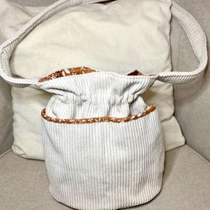 Könnte beinhalten: Ein cremefarbener Cord-Eimerbeutel mit passendem Schulterriemen. Der Innenraum und die Zierleiste weisen ein braunes Blumenmuster auf. Die Tasche steht auf einer neutral getönten Oberfläche.