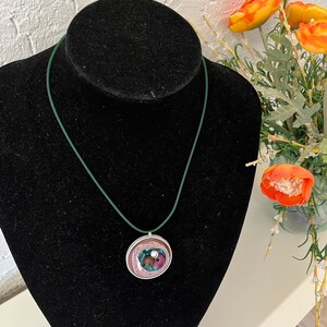 Könnte beinhalten: Eine Halskette mit grünem Band und einem runden Anhänger. Der Anhänger besteht aus übereinanderliegenden Papierkreisen in den Farben Rosa, Braun und Grün. In der Mitte des Anhängers ist eine kleine weiße Perle zu sehen.