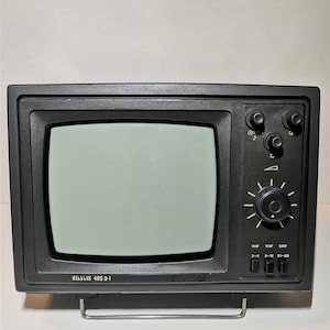 Puede incluir: Un pequeño televisor negro vintage con una pantalla rectangular. El aparato tiene varios botones y diales en el lado derecho, incluido un selector de canales. El texto "Silkkis 405 D-1" está impreso en la parte inferior de la pantalla.