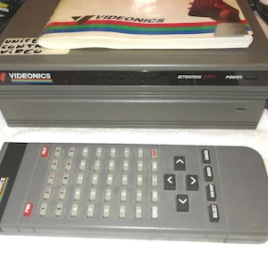 Peut inclure: Unité de contrôle vidéo grise Videonics avec une télécommande assortie. La télécommande comporte un clavier et des boutons de fonction. Un manuel repose sur l'unité. Le mot "VIDEONICS" est imprimé sur l'unité.
