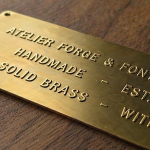Peut inclure: Plaque rectangulaire en laiton avec le texte "ATELIER FORGE & FONT - AMSTERDAM, HANDMADE, SOLID BRASS, EST. 2026, WITH CARE." La plaque a des coins arrondis et est montée sur une surface en bois foncé.