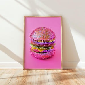 Op de afbeelding: Een ingelijste print met een glinsterende hamburger op een roze achtergrond. De hamburger heeft een roze, met pailletten bedekt broodje, met lagen sla, kaas en een patty. De lijst is lichtbruin.