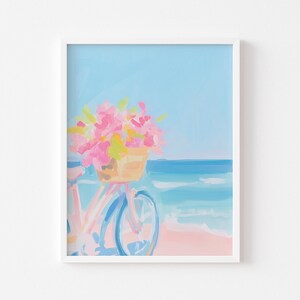 May include: A framed art print depicting a beach scene. A bicycle with a basket of pink and yellow flowers is on a pink sandy beach. The ocean is blue, and the sky is a light blue. The artwork is in a white frame.