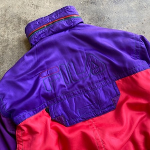 May include: A vintage Fila jacket in a vibrant colour block design. The jacket features a purple upper section with the Fila logo embroidered in green and red. The lower section is bright red, and the collar has a green, red, and yellow stripe.