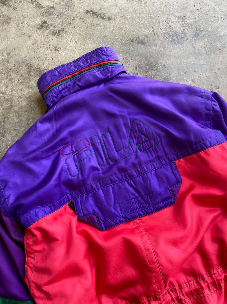May include: A vintage Fila jacket in a vibrant colour block design. The jacket features a purple upper section with the Fila logo embroidered in green and red. The lower section is bright red, and the collar has a green, red, and yellow stripe.