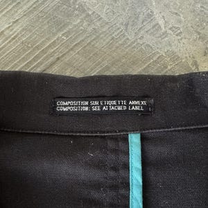 May include: A black garment featuring a rectangular fabric label with the white text COMPOSITION SUR ETIQUETTE ANNEXE and COMPOSITION: SEE ATTACHED LABEL. A vertical teal fabric trim runs downward from the center of the black collar seam.