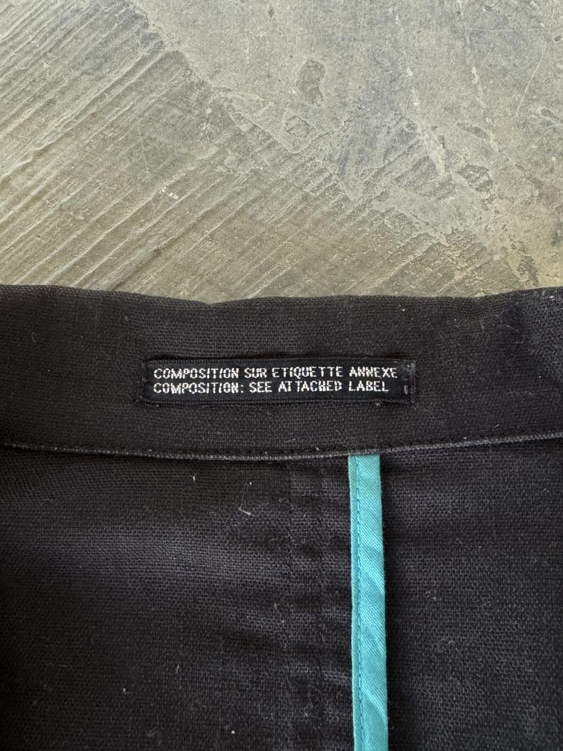 May include: A black garment featuring a rectangular fabric label with the white text COMPOSITION SUR ETIQUETTE ANNEXE and COMPOSITION: SEE ATTACHED LABEL. A vertical teal fabric trim runs downward from the center of the black collar seam.