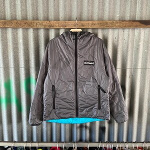 May include: A gray quilted jacket with a hood and a black zipper. The jacket has two side pockets with black zippers and a small black and white logo on the chest. The inside of the jacket is a bright blue color.