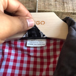 May include: A close-up of a jacket's interior, revealing a red and white checkered pattern lining. The label reads "Made in Japan Fabriqu&eacute; au Japon COMME des GAR&Ccedil;ONS". The jacket is hanging on a wooden hanger.
