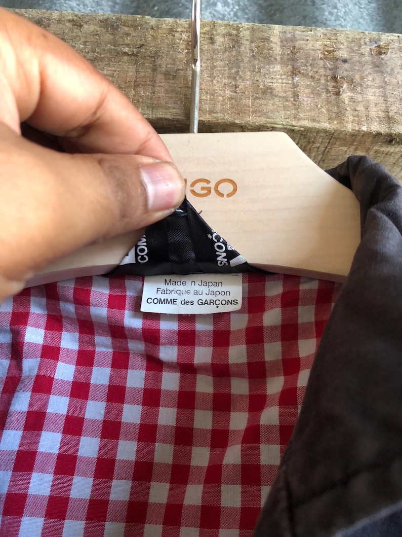 May include: A close-up of a jacket's interior, revealing a red and white checkered pattern lining. The label reads "Made in Japan Fabriqu&eacute; au Japon COMME des GAR&Ccedil;ONS". The jacket is hanging on a wooden hanger.