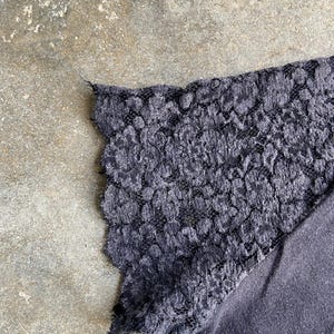 Pu&ograve; includere: Primo piano di un top grigio scuro con una manica in pizzo. Il pizzo ha un motivo floreale e un bordo smerlato. Il tessuto del top sembra essere un morbido materiale a maglia.