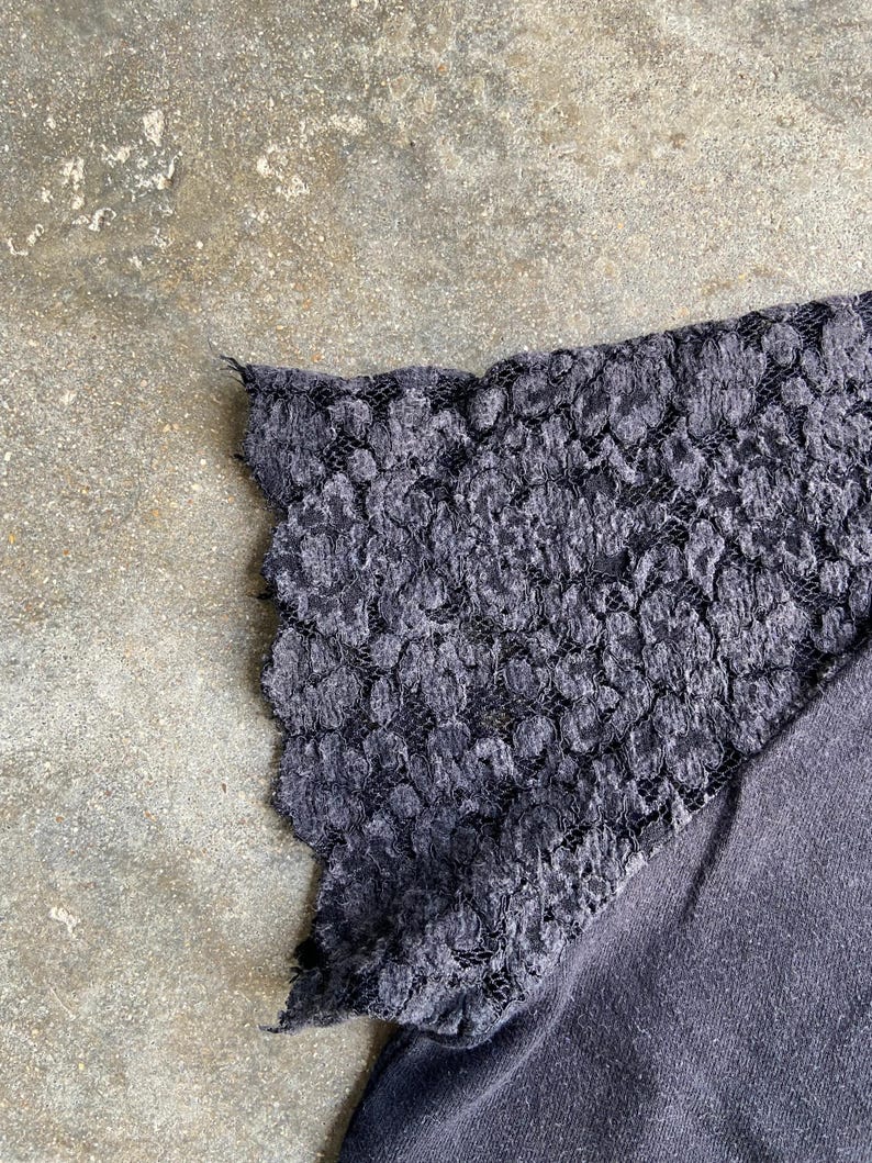 Pu&ograve; includere: Primo piano di un top grigio scuro con una manica in pizzo. Il pizzo ha un motivo floreale e un bordo smerlato. Il tessuto del top sembra essere un morbido materiale a maglia.