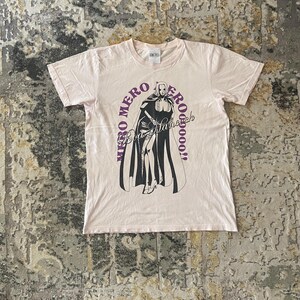 以下が含まれることがあります： 薄いピンク色のTシャツ。黒と白のイラストで、マントを着たキャラクターが描かれています。「Miso Mero Eroooooo! Boa Hancock」という文字が紫でキャラクターの周りにプリントされています。