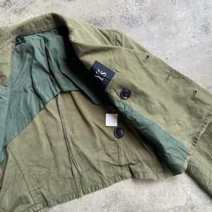 May include: Olive green jacket with a contrasting dark green panel and lining. Features black buttons and a small black label with white text. The jacket has a classic design.
