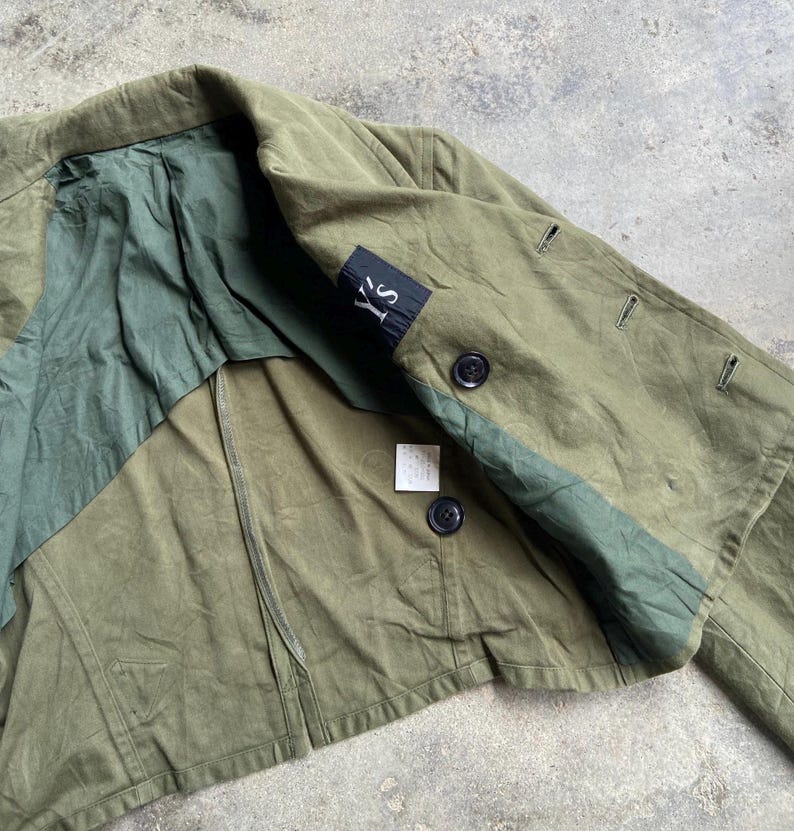 May include: Olive green jacket with a contrasting dark green panel and lining. Features black buttons and a small black label with white text. The jacket has a classic design.