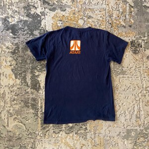 Puede incluir: Camiseta azul marino con el logo de Atari en naranja y blanco. El logo es un diseño estilizado con la palabra "ATARI" debajo. La camiseta está hecha de un material suave y tiene mangas cortas.