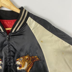 Puede incluir: Chaqueta bomber de sat&eacute;n negro con bordado de tigre. La chaqueta tiene un panel color crema en la manga y un forro rojo. El cuello y los pu&ntilde;os son negros con rayas crema. La chaqueta est&aacute; colgada en una percha de madera.