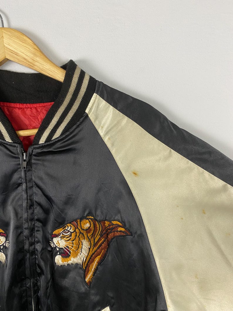 Puede incluir: Chaqueta bomber de sat&eacute;n negro con bordado de tigre. La chaqueta tiene un panel color crema en la manga y un forro rojo. El cuello y los pu&ntilde;os son negros con rayas crema. La chaqueta est&aacute; colgada en una percha de madera.
