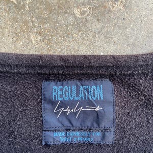 Pu&ograve; includere: Primo piano di una felpa grigio scuro con un'etichetta blu navy. L'etichetta recita "REGULATION" in testo azzurro, con una firma e la scritta "MADE EXPRESSLY FOR YOUTH & PEOPLE."