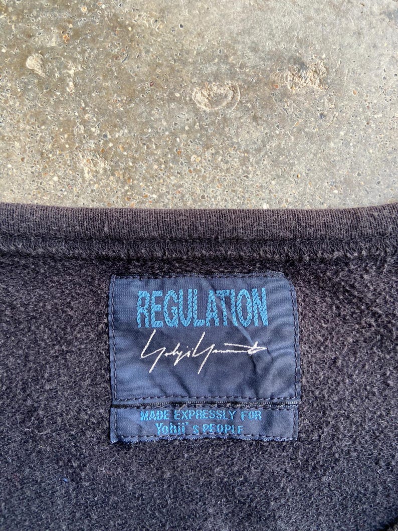 Pu&ograve; includere: Primo piano di una felpa grigio scuro con un'etichetta blu navy. L'etichetta recita "REGULATION" in testo azzurro, con una firma e la scritta "MADE EXPRESSLY FOR YOUTH & PEOPLE."