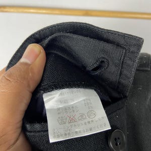 May include: Close-up of a black garment with a label attached. The label displays text in Japanese, including size and fabric composition. A black button is visible on the garment.