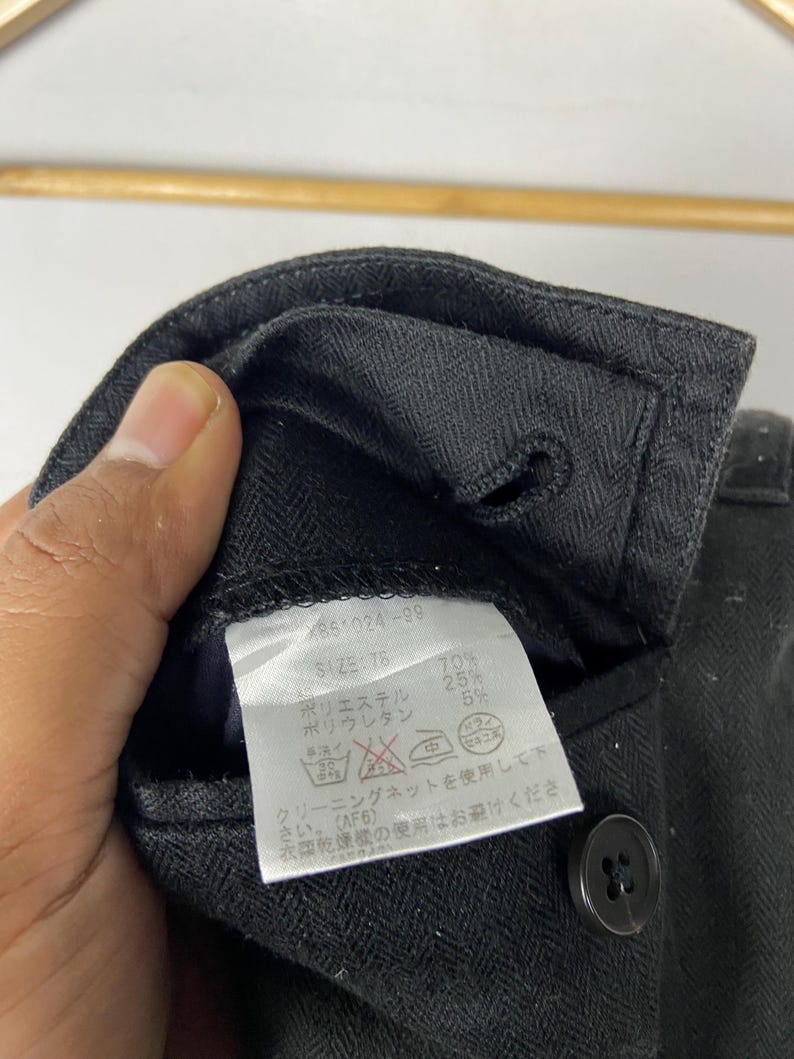 May include: Close-up of a black garment with a label attached. The label displays text in Japanese, including size and fabric composition. A black button is visible on the garment.