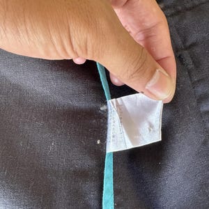 May include: A black fabric garment featuring a white care instruction label with Japanese text and a thin, vertical teal trim strip running along the seam. The material has a textured, woven appearance with visible stitching details.