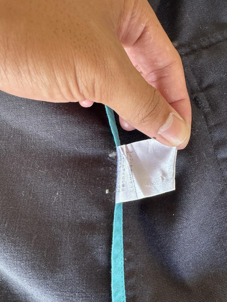 May include: A black fabric garment featuring a white care instruction label with Japanese text and a thin, vertical teal trim strip running along the seam. The material has a textured, woven appearance with visible stitching details.