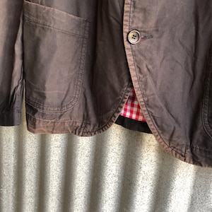 May include: Close-up of a brown blazer with two buttons and a pocket. The blazer has a red and white checkered lining. The fabric appears to be a sturdy cotton or similar material.