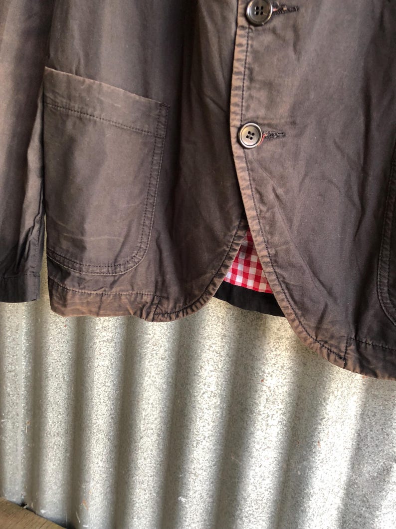 May include: Close-up of a brown blazer with two buttons and a pocket. The blazer has a red and white checkered lining. The fabric appears to be a sturdy cotton or similar material.
