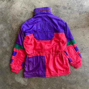 May include: A vintage purple, pink, and green jacket with the word "FILA" on the back. The jacket has a high collar and a drawstring waist. The sleeves have green and purple stripes. The jacket is made of a shiny, possibly nylon, material.