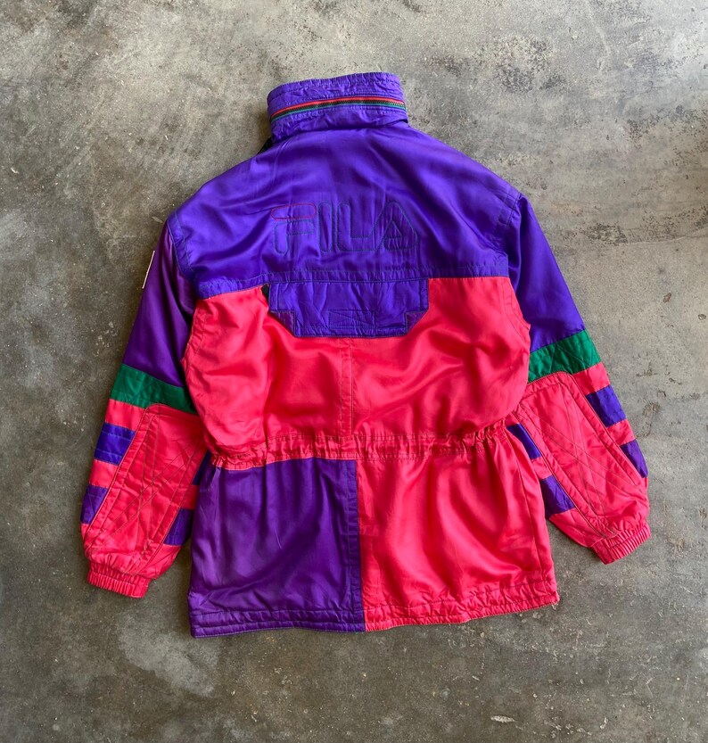 May include: A vintage purple, pink, and green jacket with the word "FILA" on the back. The jacket has a high collar and a drawstring waist. The sleeves have green and purple stripes. The jacket is made of a shiny, possibly nylon, material.