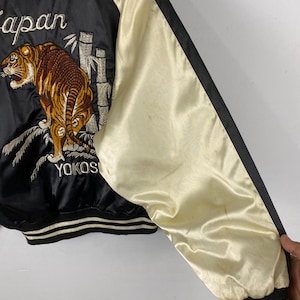 Puede incluir: Chaqueta bomber de sat&eacute;n negro con mangas color crema. La chaqueta presenta un dise&ntilde;o bordado de tigre y bamb&uacute; con las palabras "Japan" y "Yokos" en blanco. Los pu&ntilde;os y el dobladillo tienen detalles a rayas blancas y negras.