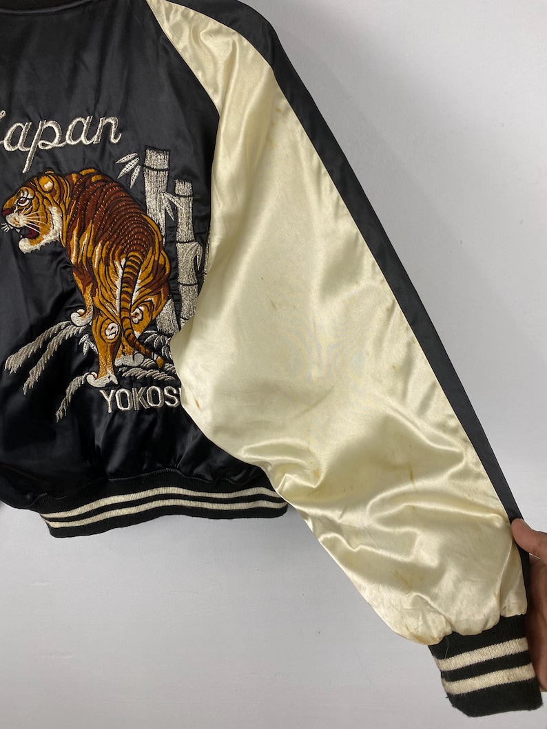Puede incluir: Chaqueta bomber de sat&eacute;n negro con mangas color crema. La chaqueta presenta un dise&ntilde;o bordado de tigre y bamb&uacute; con las palabras "Japan" y "Yokos" en blanco. Los pu&ntilde;os y el dobladillo tienen detalles a rayas blancas y negras.