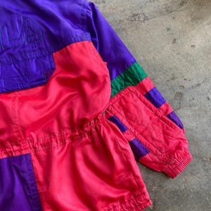May include: A vintage colour-blocked jacket in purple, pink, and green. The jacket features the brand name "FILA" embroidered in purple on the upper left chest. The jacket has a zip and elastic cuffs.