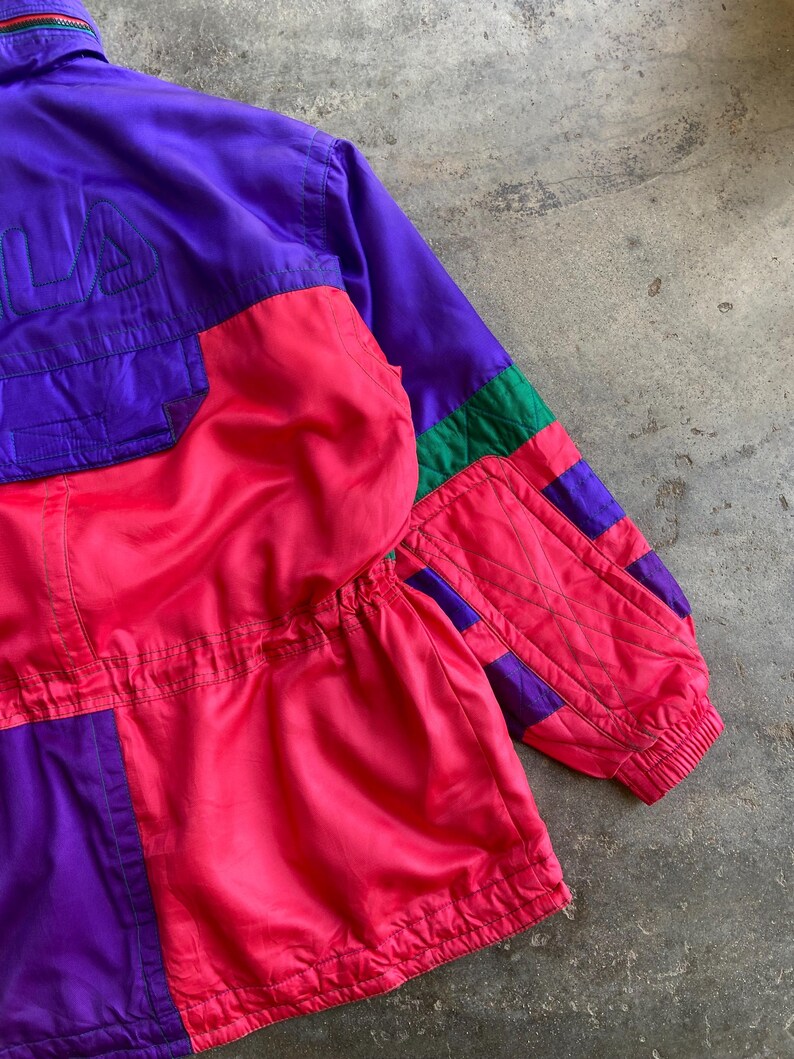 May include: A vintage colour-blocked jacket in purple, pink, and green. The jacket features the brand name "FILA" embroidered in purple on the upper left chest. The jacket has a zip and elastic cuffs.