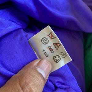 May include: A white care label with symbols and the text "Y-TK03" is held against a purple fabric. The label displays various symbols indicating washing and care instructions for the garment.
