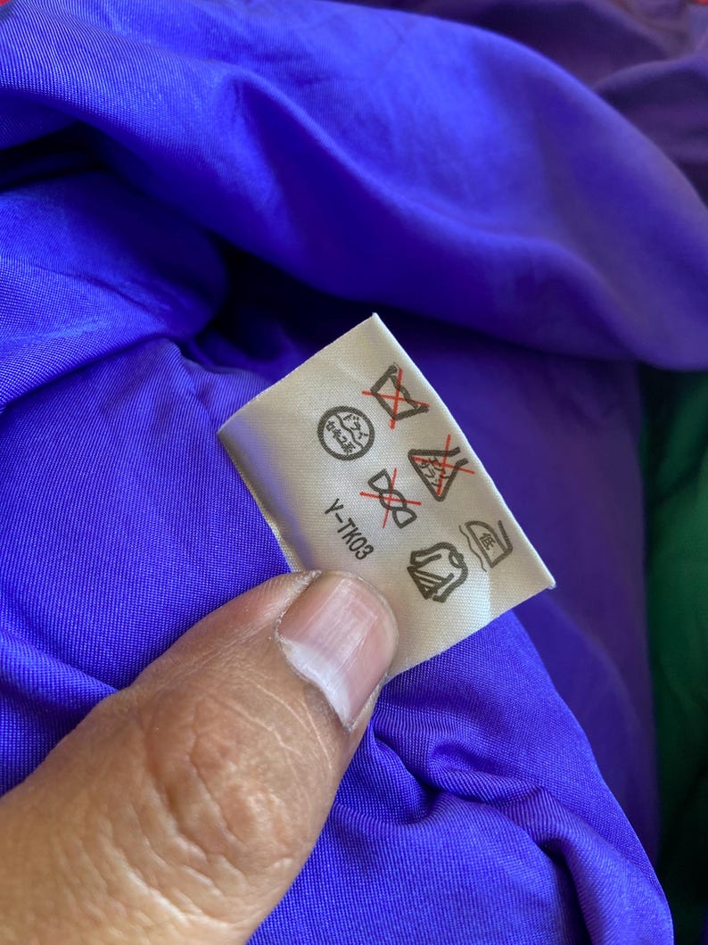 May include: A white care label with symbols and the text "Y-TK03" is held against a purple fabric. The label displays various symbols indicating washing and care instructions for the garment.