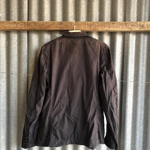 May include: A dark brown jacket hanging on a wooden beam. The jacket has long sleeves, a collar, and buttons on the cuffs. The jacket is made of a lightweight material and appears to be slightly wrinkled.