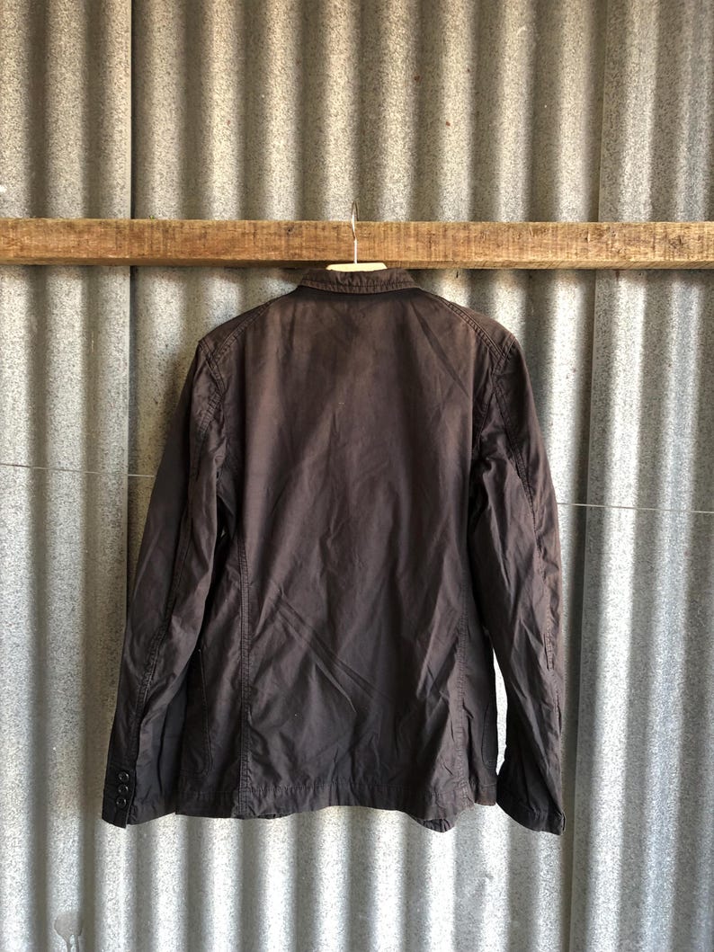 May include: A dark brown jacket hanging on a wooden beam. The jacket has long sleeves, a collar, and buttons on the cuffs. The jacket is made of a lightweight material and appears to be slightly wrinkled.