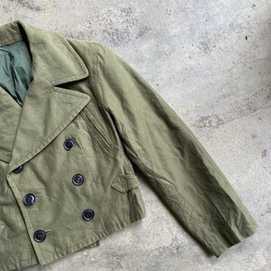 May include: An olive green double-breasted jacket with black buttons. The jacket has a notched collar, two front pockets, and a green lining. The sleeves are long and the fabric appears to be a durable cotton or blend.