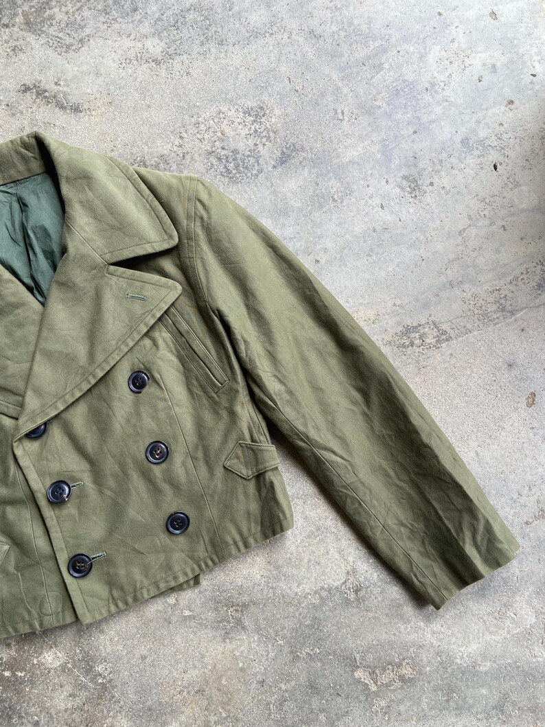 May include: An olive green double-breasted jacket with black buttons. The jacket has a notched collar, two front pockets, and a green lining. The sleeves are long and the fabric appears to be a durable cotton or blend.