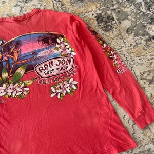 May include: Coral long-sleeve shirt featuring a tropical graphic with the text "RON JON SURF SHOP ONE AND ONLY". The design includes a beach scene with palm trees, flowers, and a surfer. The sleeve also has a smaller "RON JON SURF SHOP" logo.