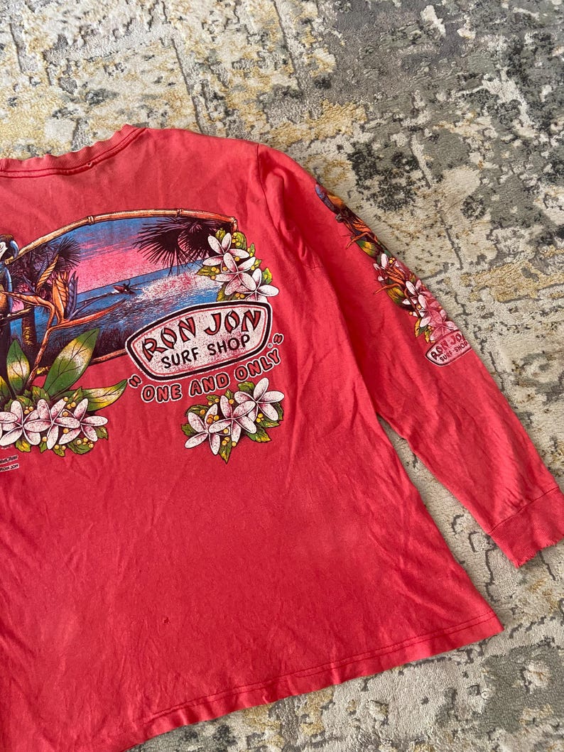 May include: Coral long-sleeve shirt featuring a tropical graphic with the text "RON JON SURF SHOP ONE AND ONLY". The design includes a beach scene with palm trees, flowers, and a surfer. The sleeve also has a smaller "RON JON SURF SHOP" logo.