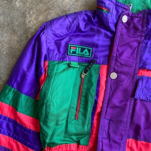 May include: A retro Fila jacket in a striking colour block design. The jacket features a purple body with green and red accents. The Fila logo is displayed on the chest. The jacket has a zip closure and multiple pockets.