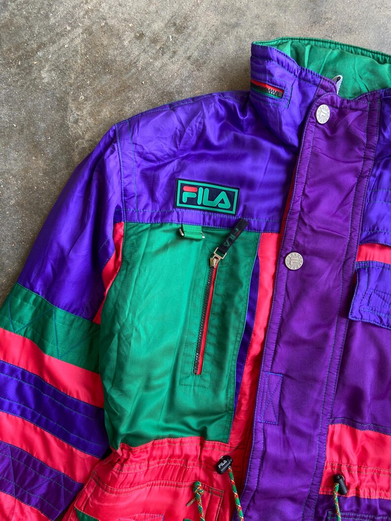 May include: A retro Fila jacket in a striking colour block design. The jacket features a purple body with green and red accents. The Fila logo is displayed on the chest. The jacket has a zip closure and multiple pockets.