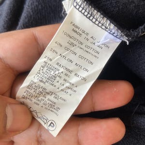Pu&ograve; includere: Un'etichetta di abbigliamento blu navy con testo in inglese e francese, che descrive la composizione del tessuto e le istruzioni per la cura. L'etichetta recita "FABRIQUE AU JAPON MADE IN JAPAN" ed elenca le percentuali di cotone, nylon e rayon.